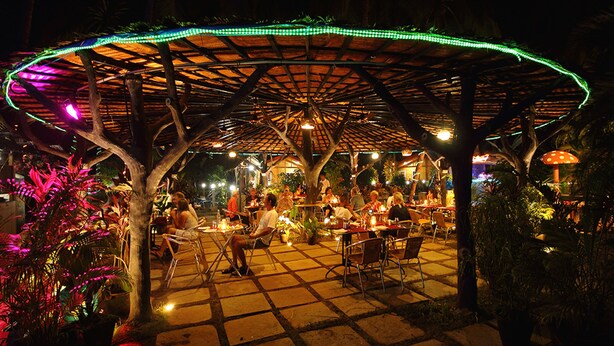 An outdoor dining area with a covered structure, tables, chairs, and ambient lighting. - Ala Goa Resort (Betalbatim)