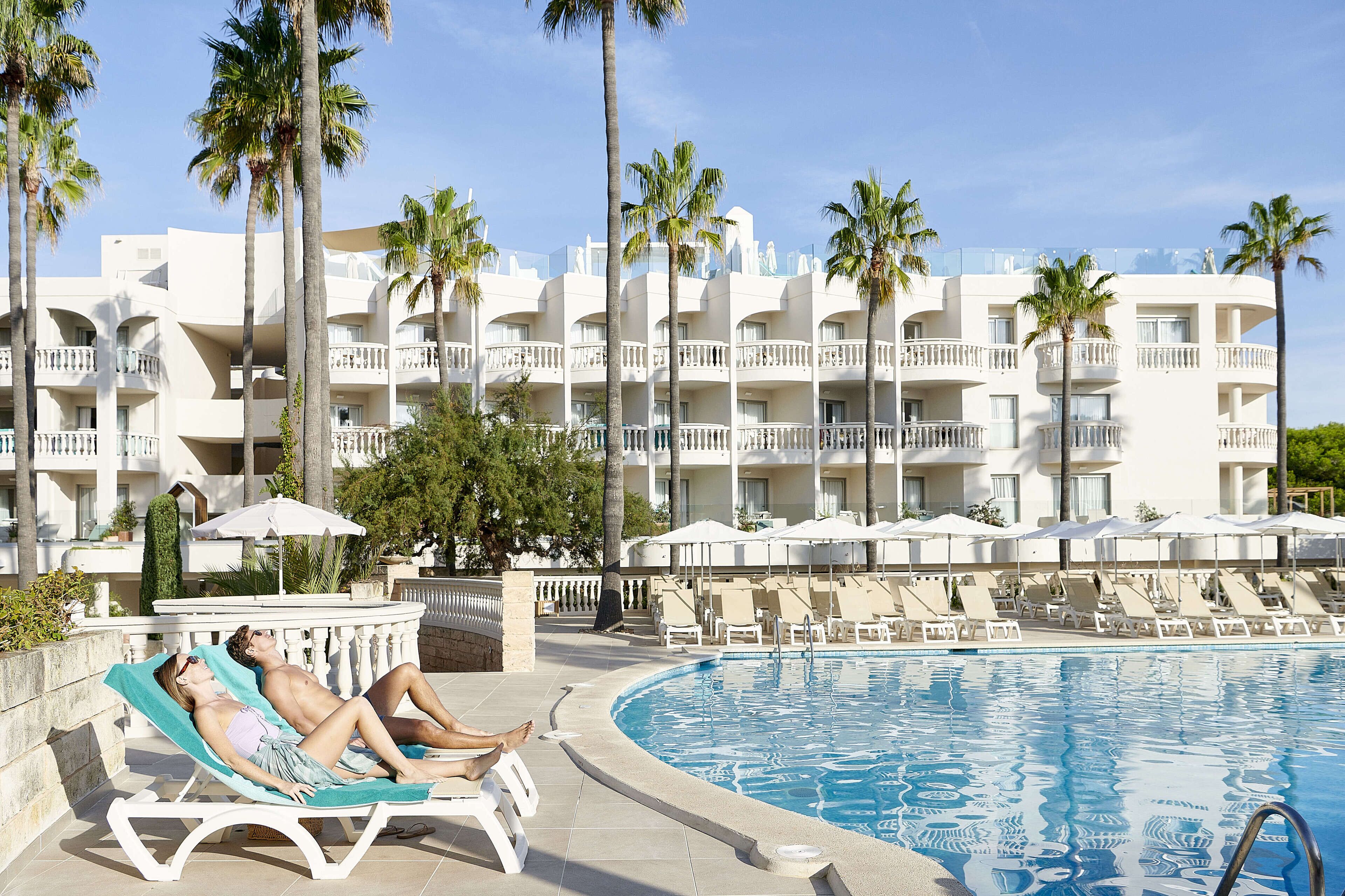 Zona de piscina con sillones de descanso y sombrillas, rodeada de palmeras y un edificio hotelero de varios pisos.