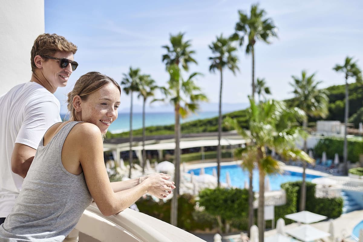 A couple on a balcony overlooking a pool with palm trees and a clear sky.