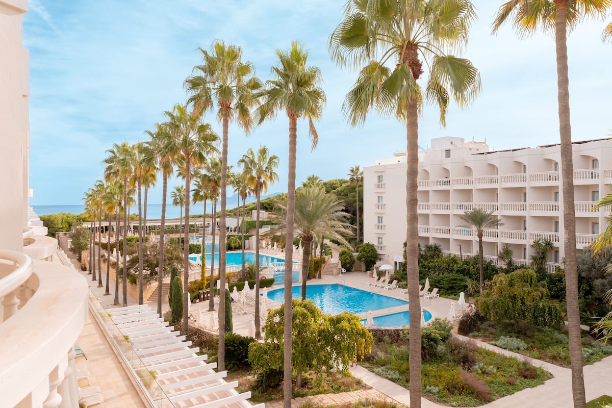 A resort with a pool surrounded by palm trees, a white building with balconies, and a clear view of the ocean.