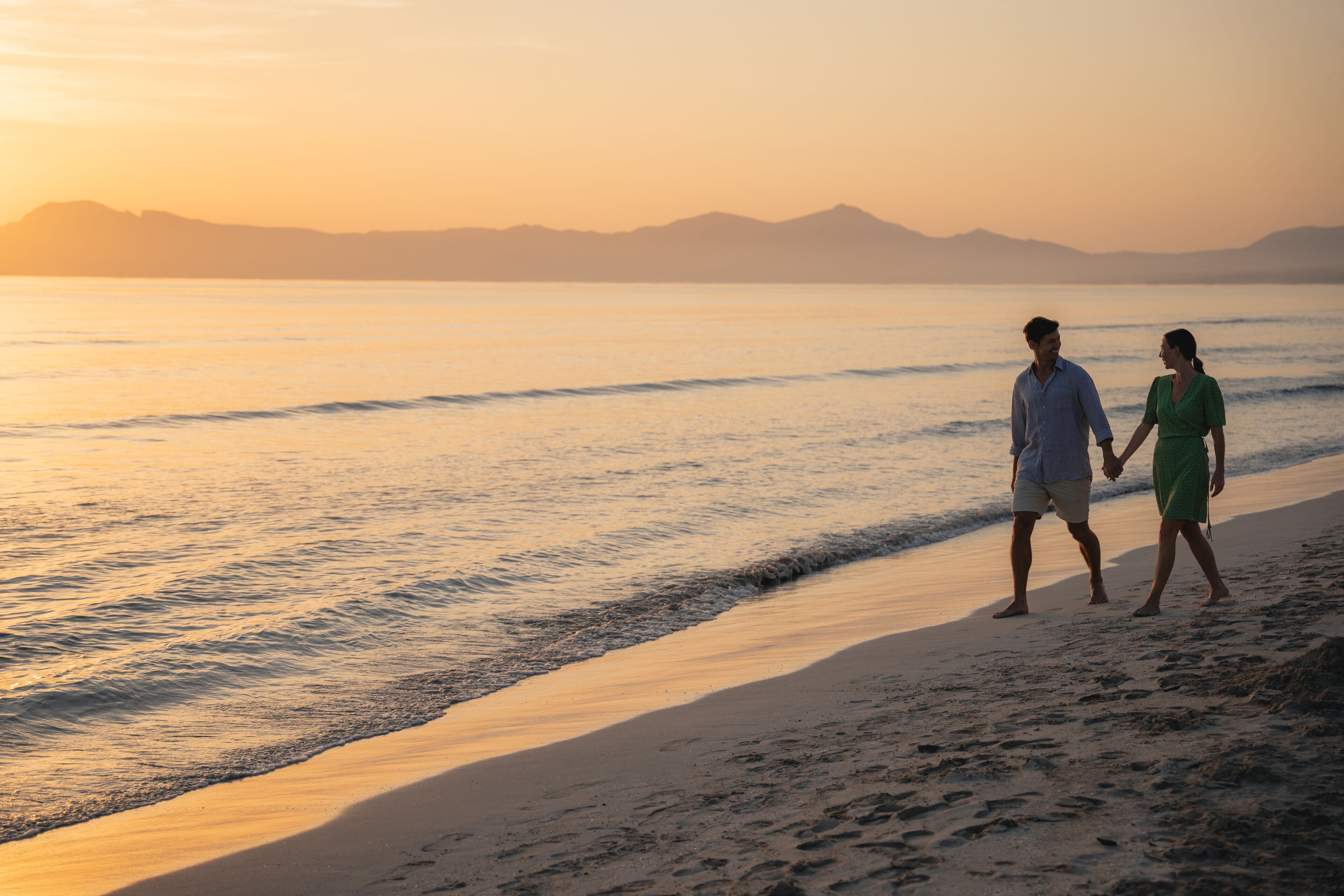 Una pareja caminando por la playa al atardecer, con montañas al fondo.