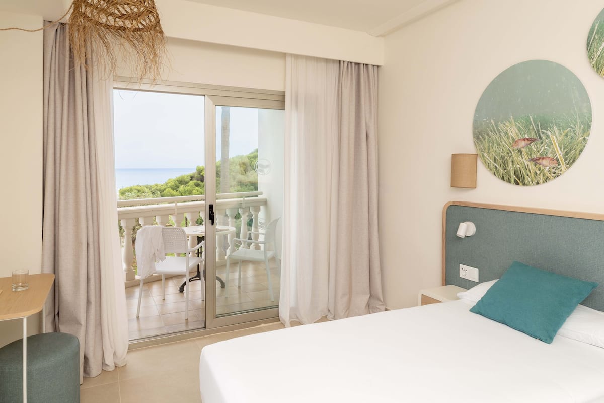 A hotel room with a balcony, a bed with a teal pillow, a round wall decoration, and a woven light fixture.