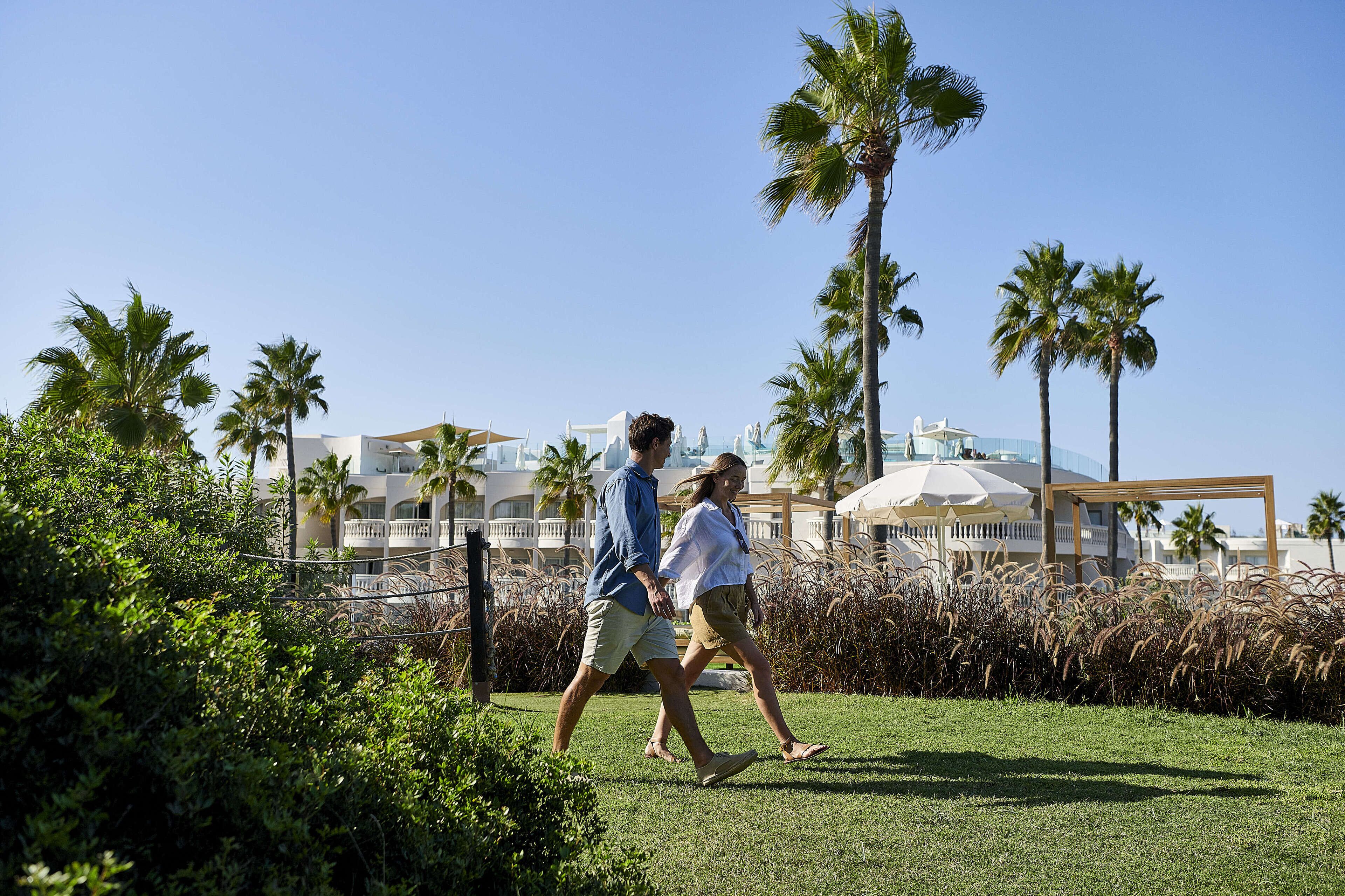 Una pareja caminando sobre un césped con palmeras y un edificio al fondo.