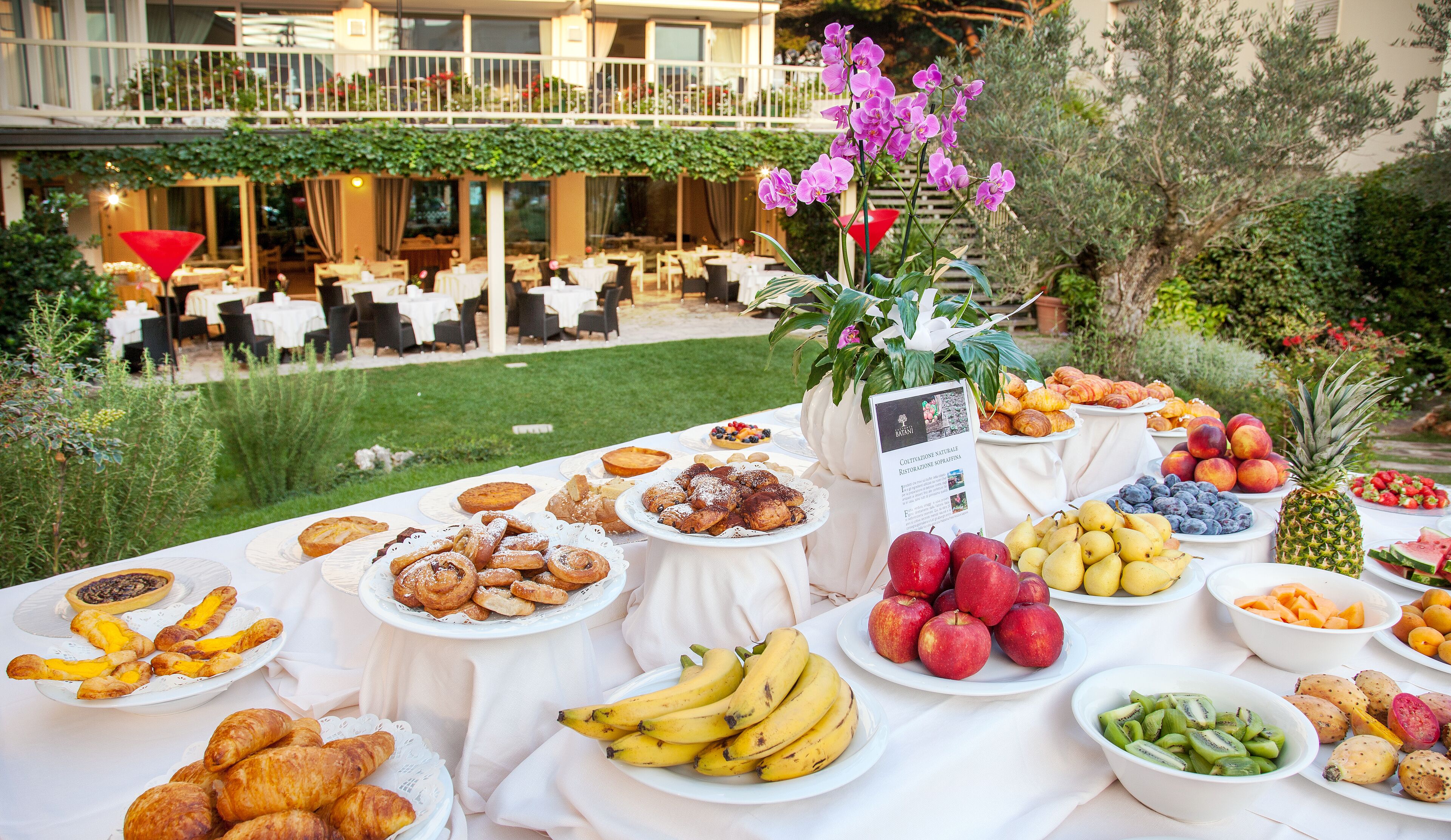 An outdoor buffet with various fruits, pastries, and a signboard.