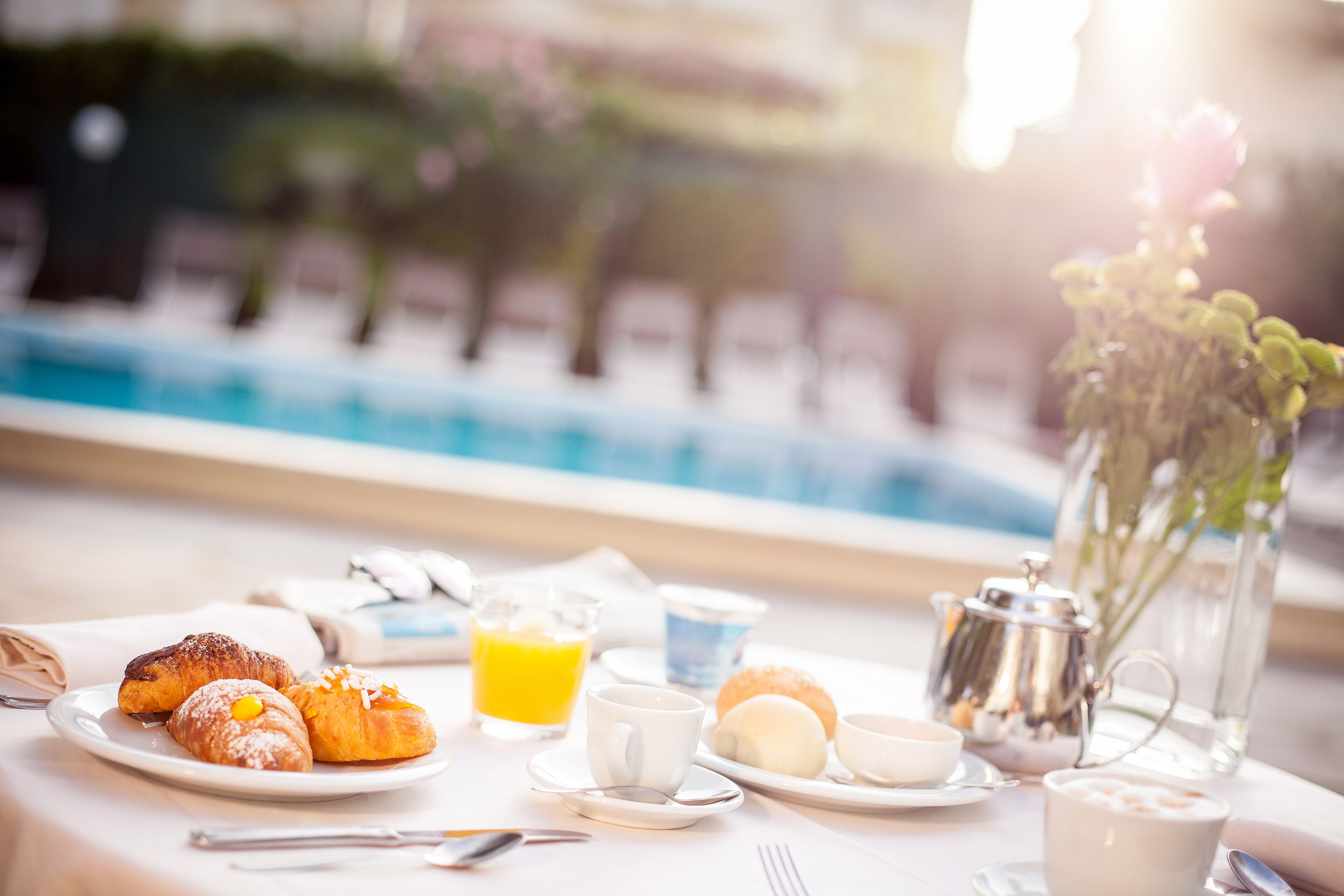 Desayuno con cruasanes, zumo de naranja, café y té en una mesa junto a una piscina.