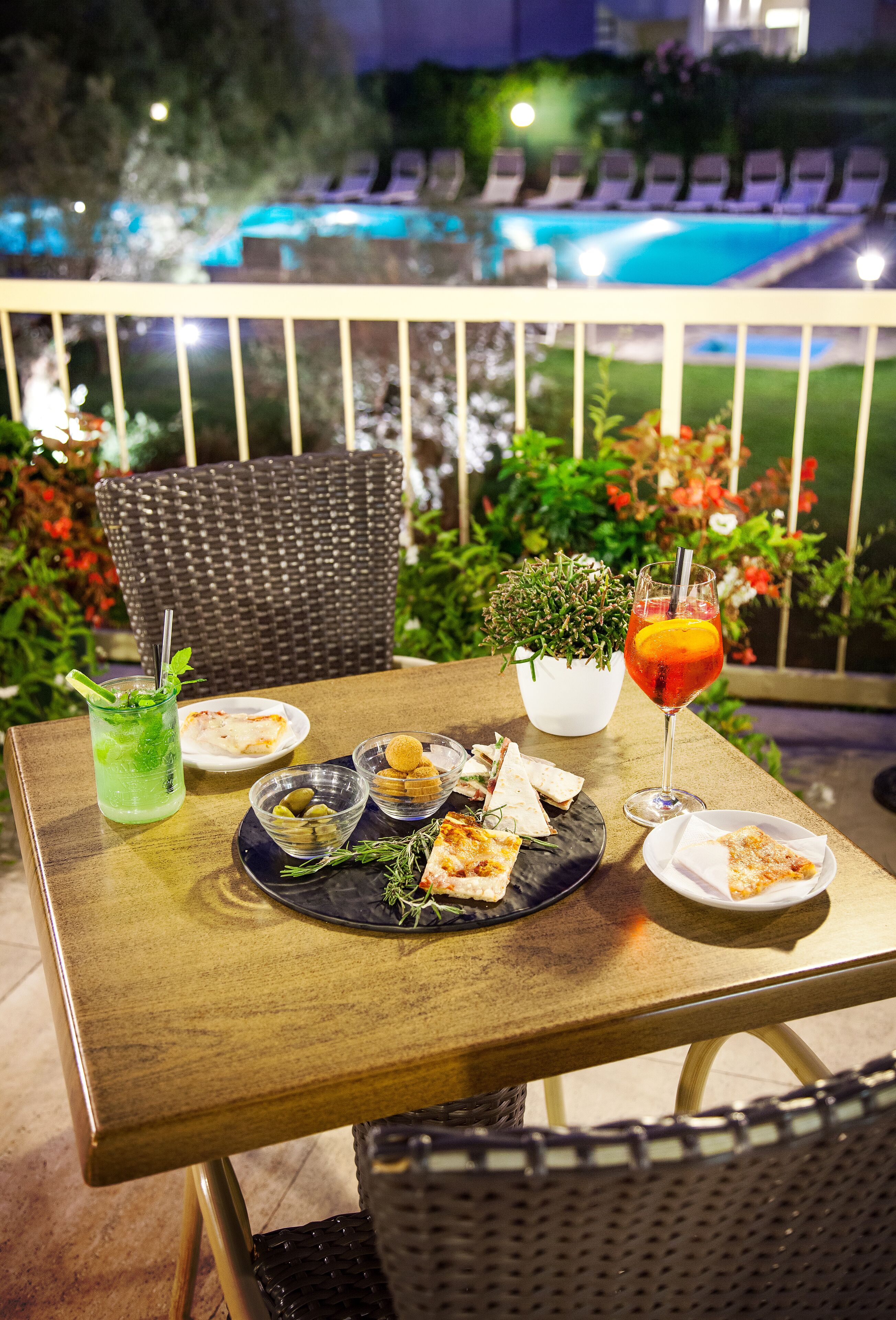 Mesa de comedor al aire libre con comida, bebidas y vistas a una piscina y sillones de descanso.