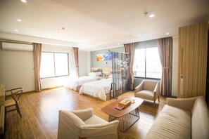 A hotel room with a bed, a sofa, a coffee table, and a chair. There is a window with curtains, a wall-mounted air conditioner, and a painting.