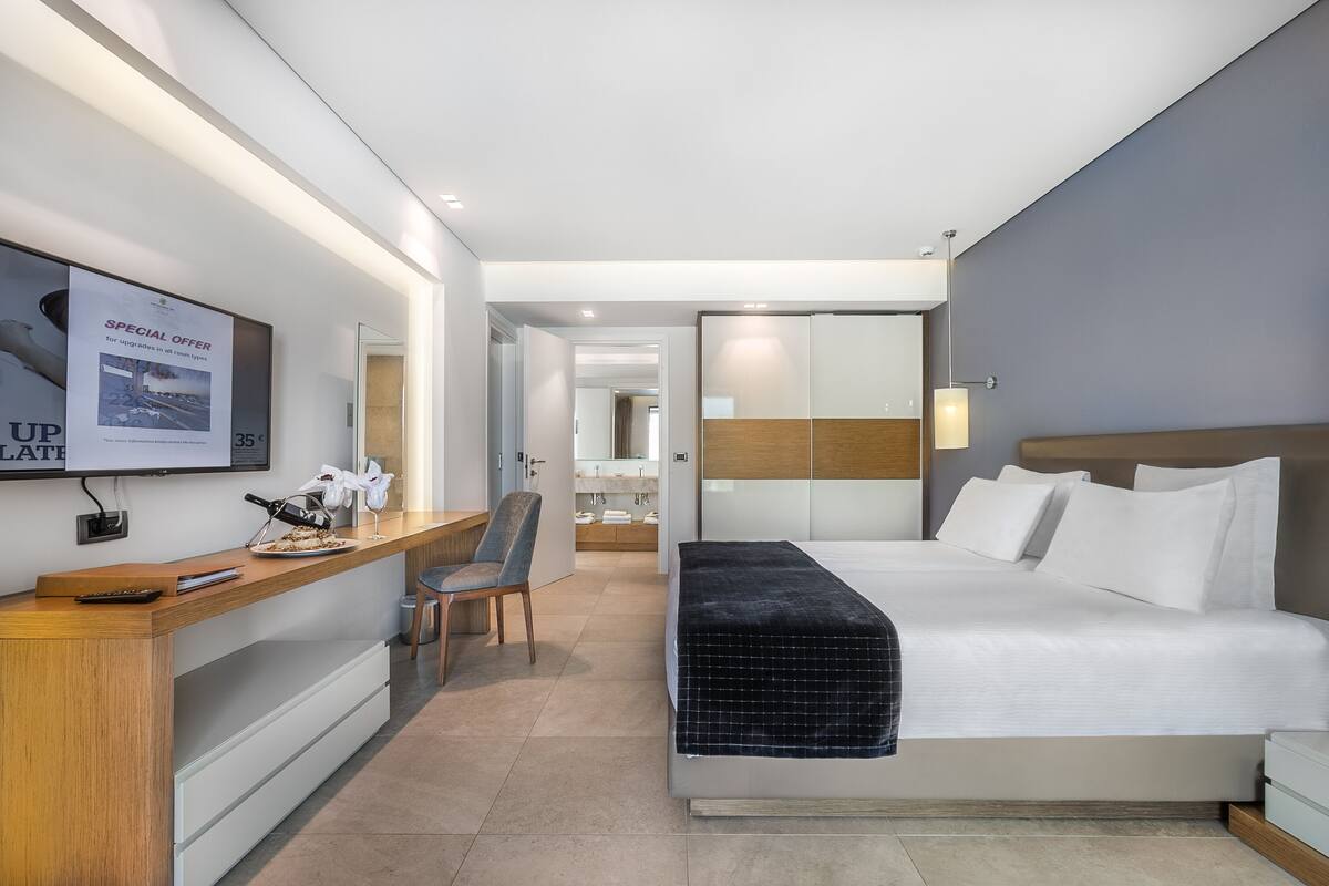 A modern hotel room with a large bed, a desk with a chair, a mirror, and a TV on the wall.