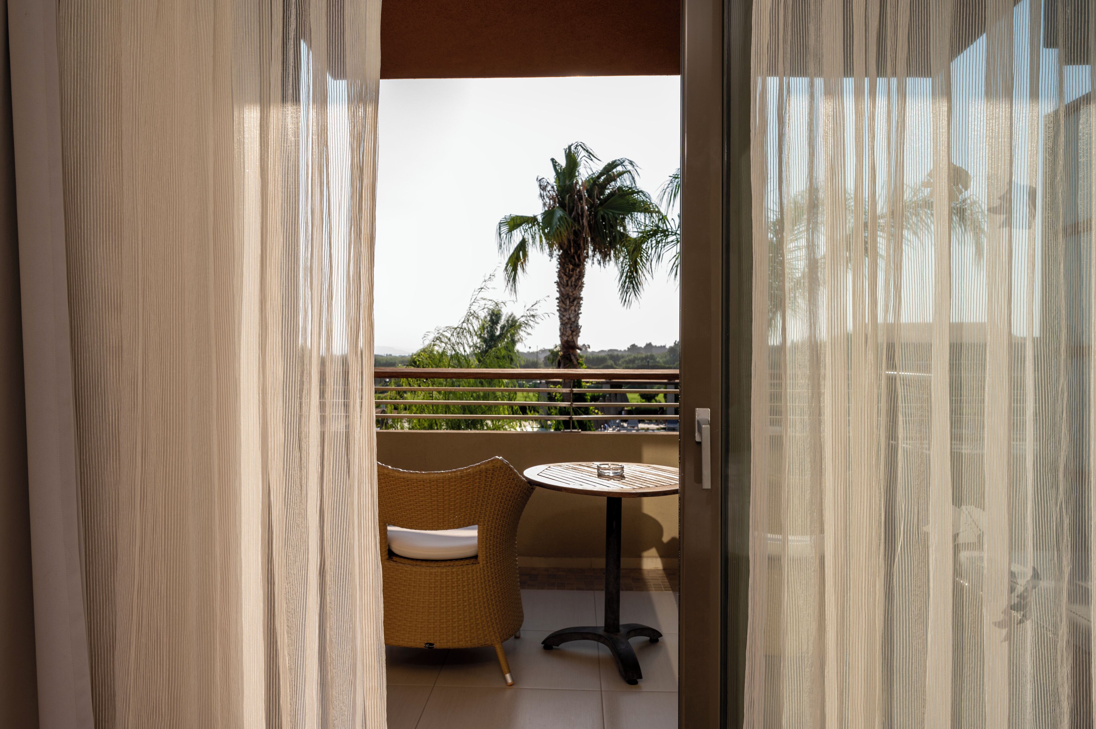 A balcony with a wicker chair and a small round table, a palm tree, and a view of greenery.