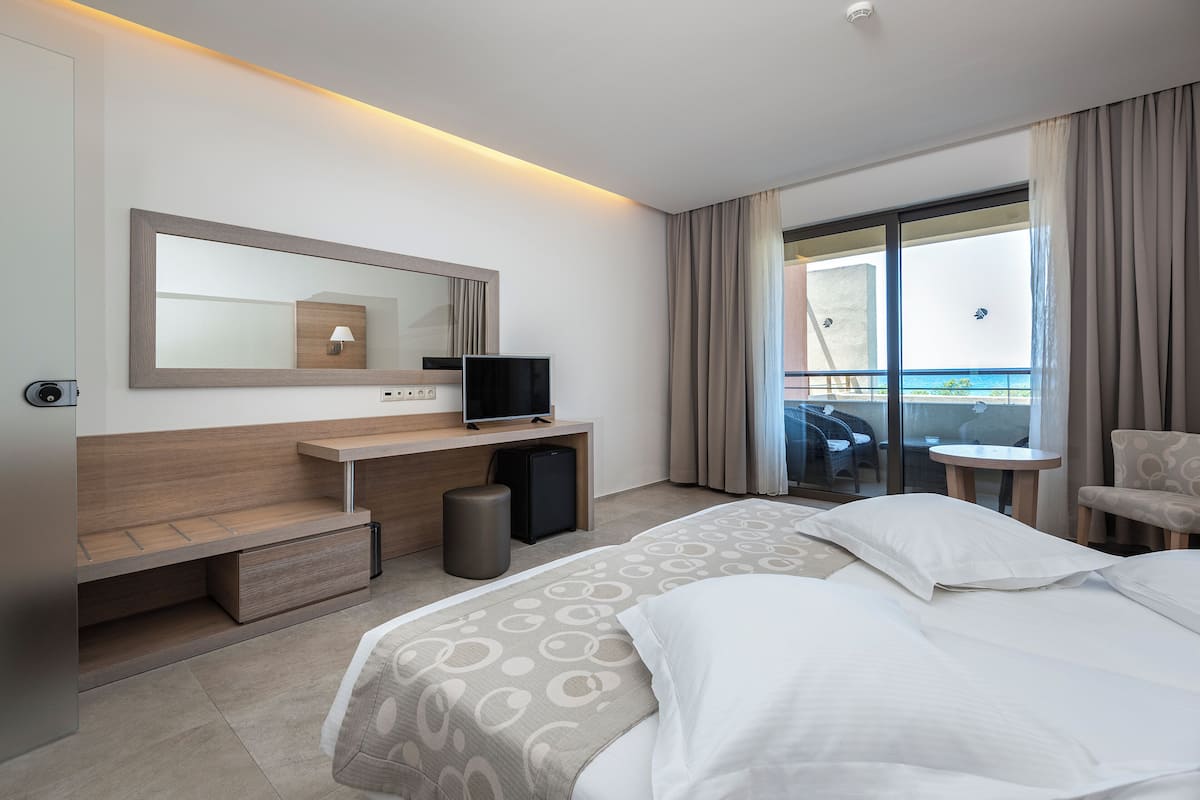 A modern hotel room with a bed, a desk with a TV, a small fridge, and a balcony with a view.