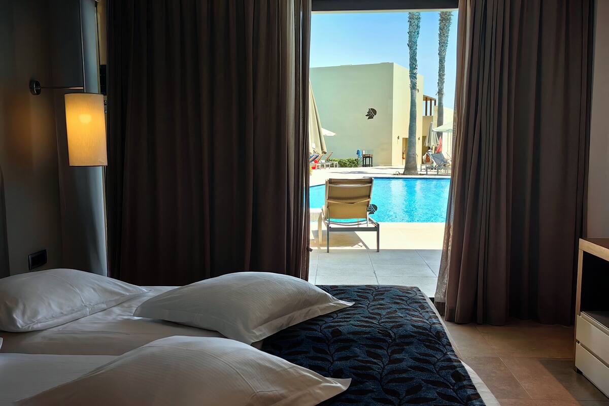 A hotel room with a bed, a view of a swimming pool, and a chair outside.