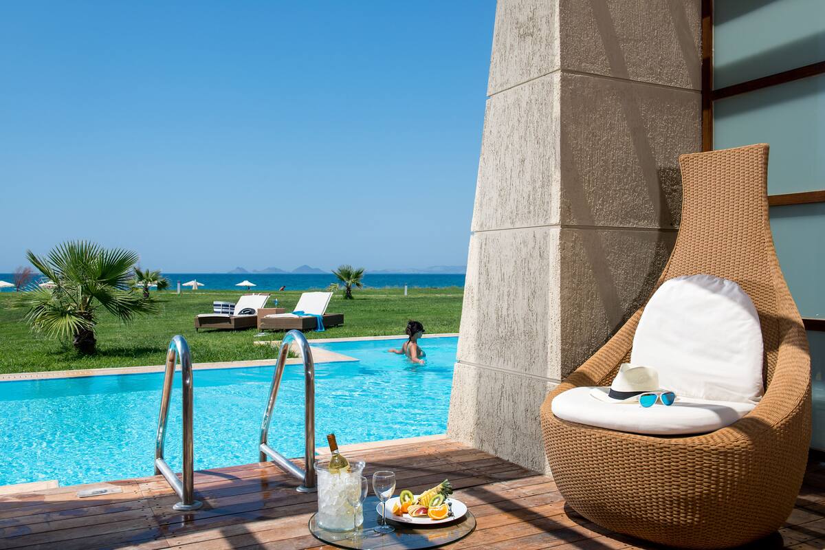 A poolside area with a wicker lounge chair, a table with a drink and fruit, and a view of the ocean and pool.