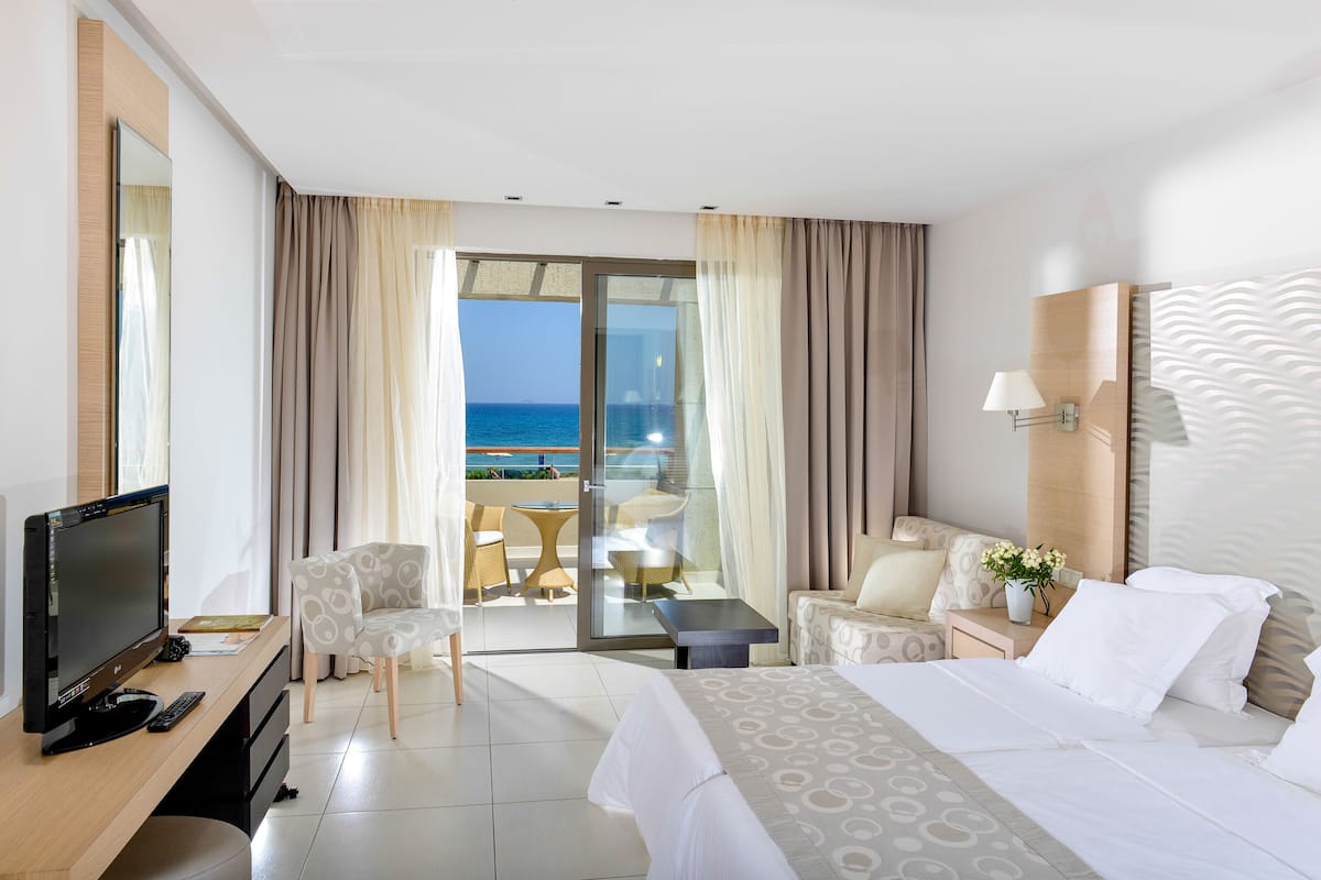 A hotel room with a bed, a desk with a TV, a chair, a small table, and a balcony with a view of the sea.