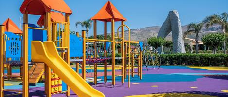 Un parque infantil con toboganes y estructuras para escalar, rodeado de un área ajardinada con palmeras y una montaña al fondo.