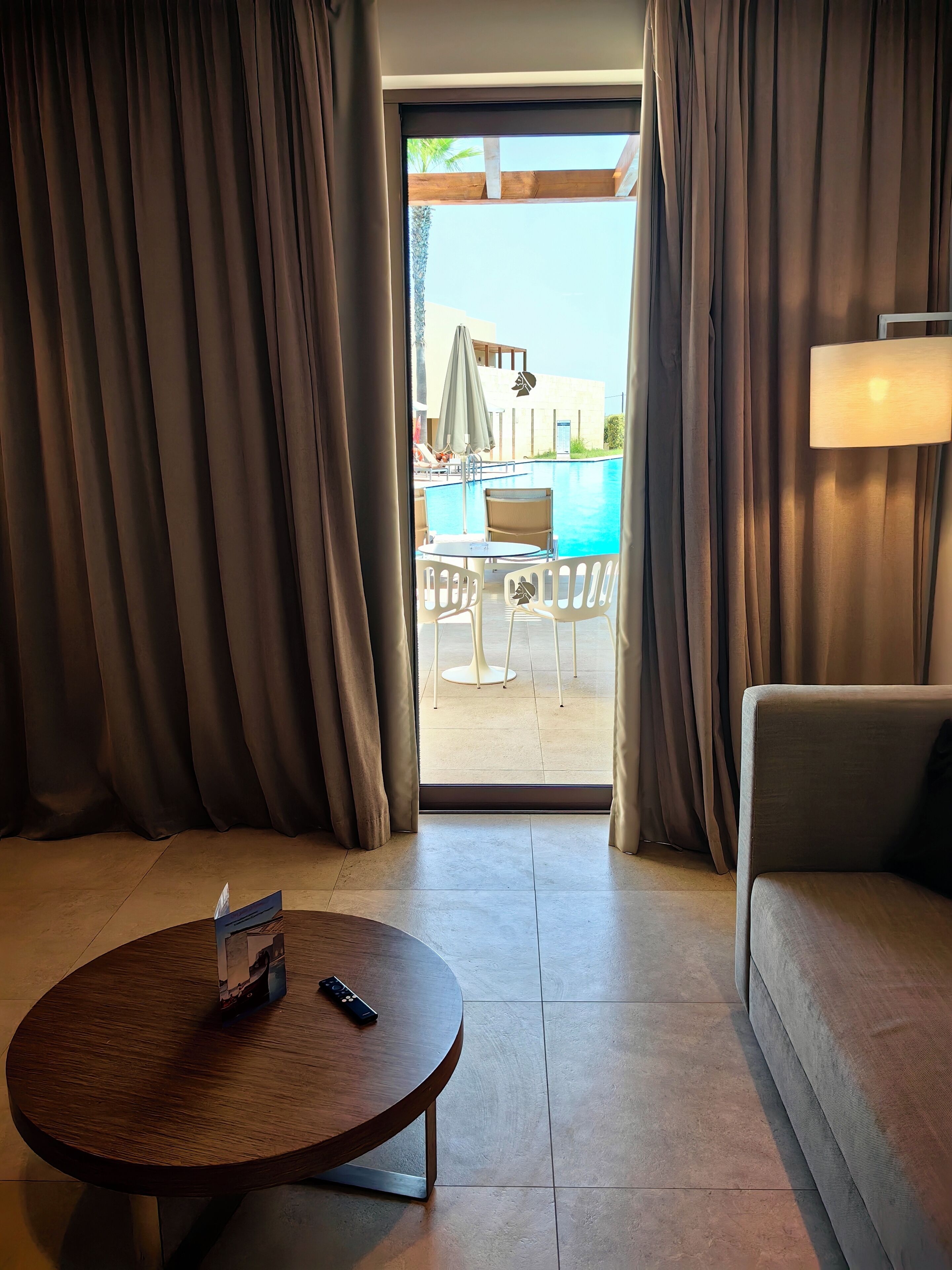 A hotel room with a view of an outdoor swimming pool, a round wooden table, a sofa, and a lamp.