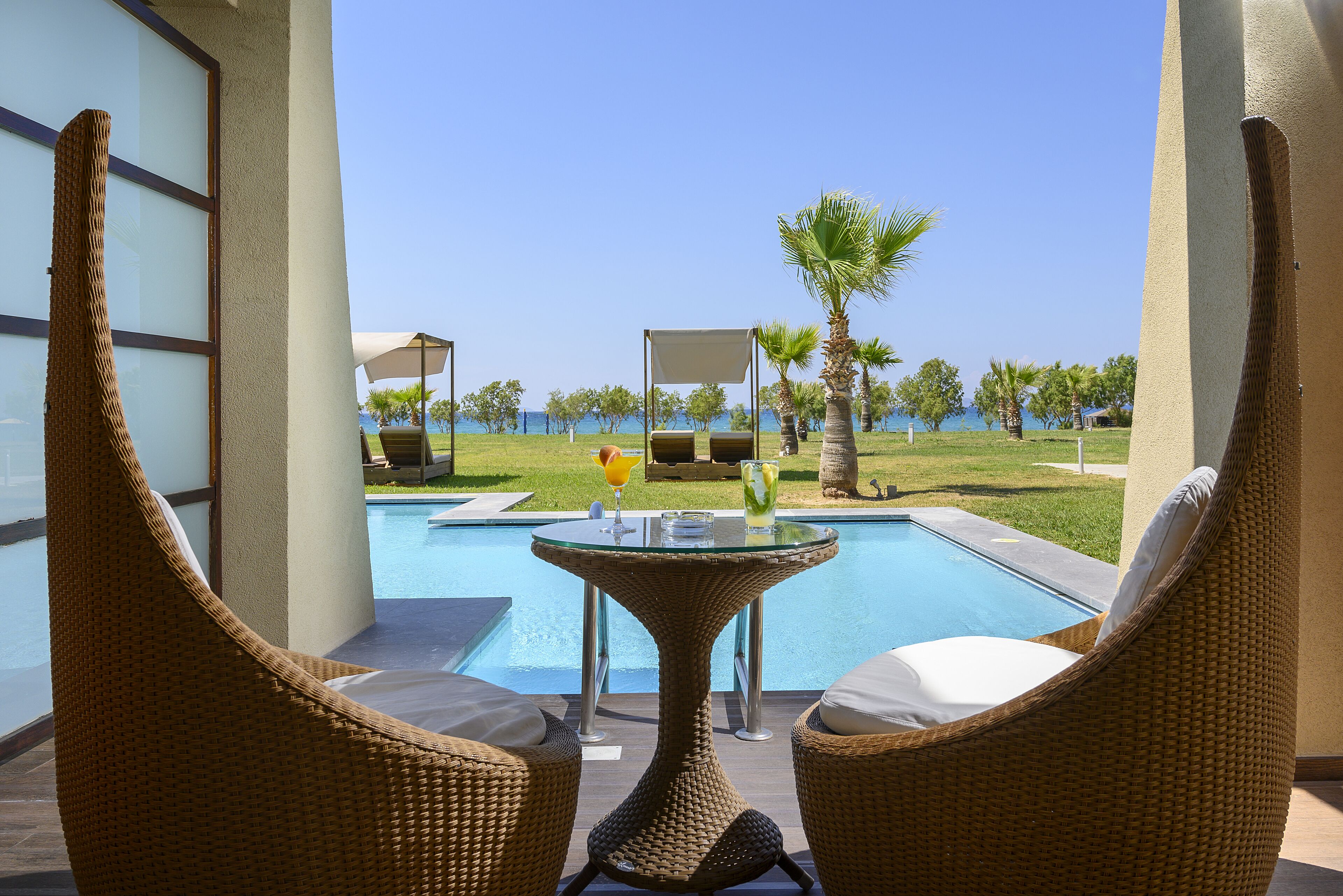 Zona junto a la piscina con sillas de mimbre y una mesa de cristal, con vistas a la piscina y al mar.