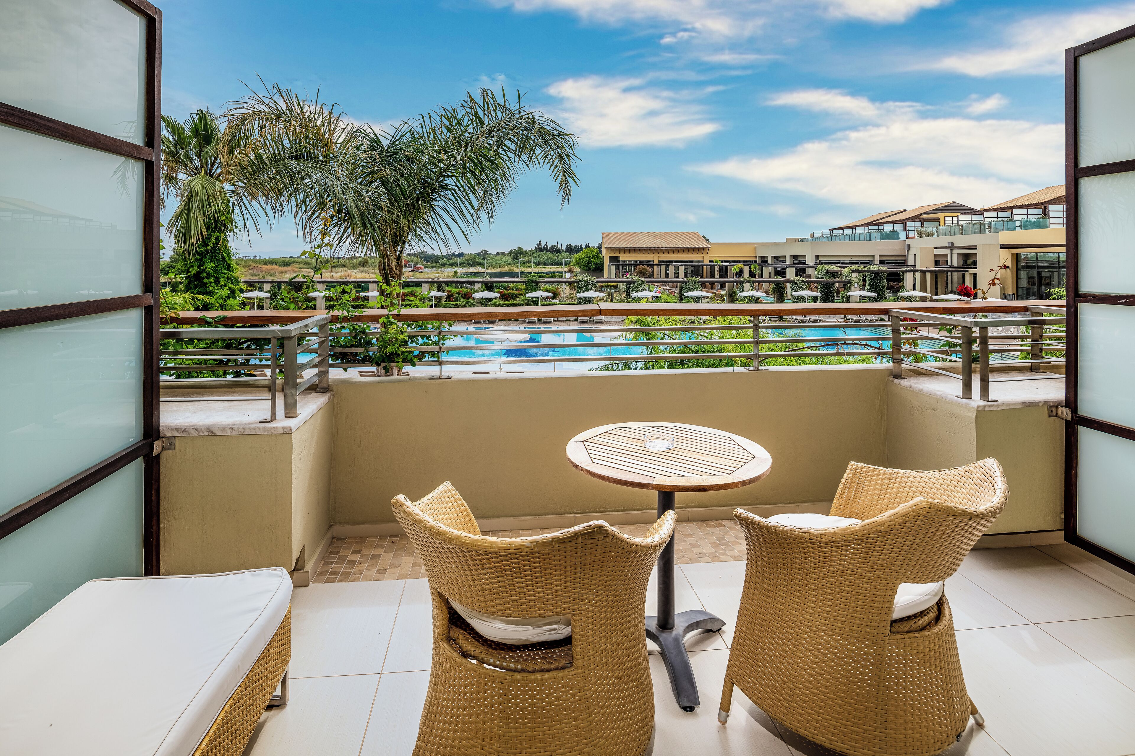 A balcony with two wicker chairs, a small round table, and a view of a swimming pool and buildings in the background.
