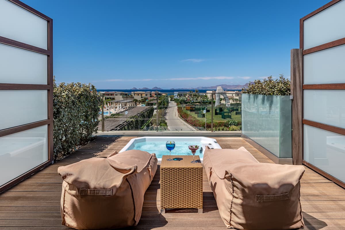 A balcony with a hot tub, two cushioned chairs, and a view of a cityscape and mountains.