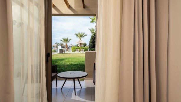 An open doorway leading to a patio with a round table and chairs, a grassy area, and a building with palm trees in the background. - Astir Odysseus Kos Resort & Spa (Kos)