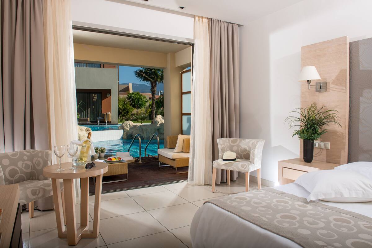A hotel room with a bed, a small table with drinks, a chair, a nightstand with a lamp, and a view of a pool and outdoor area.