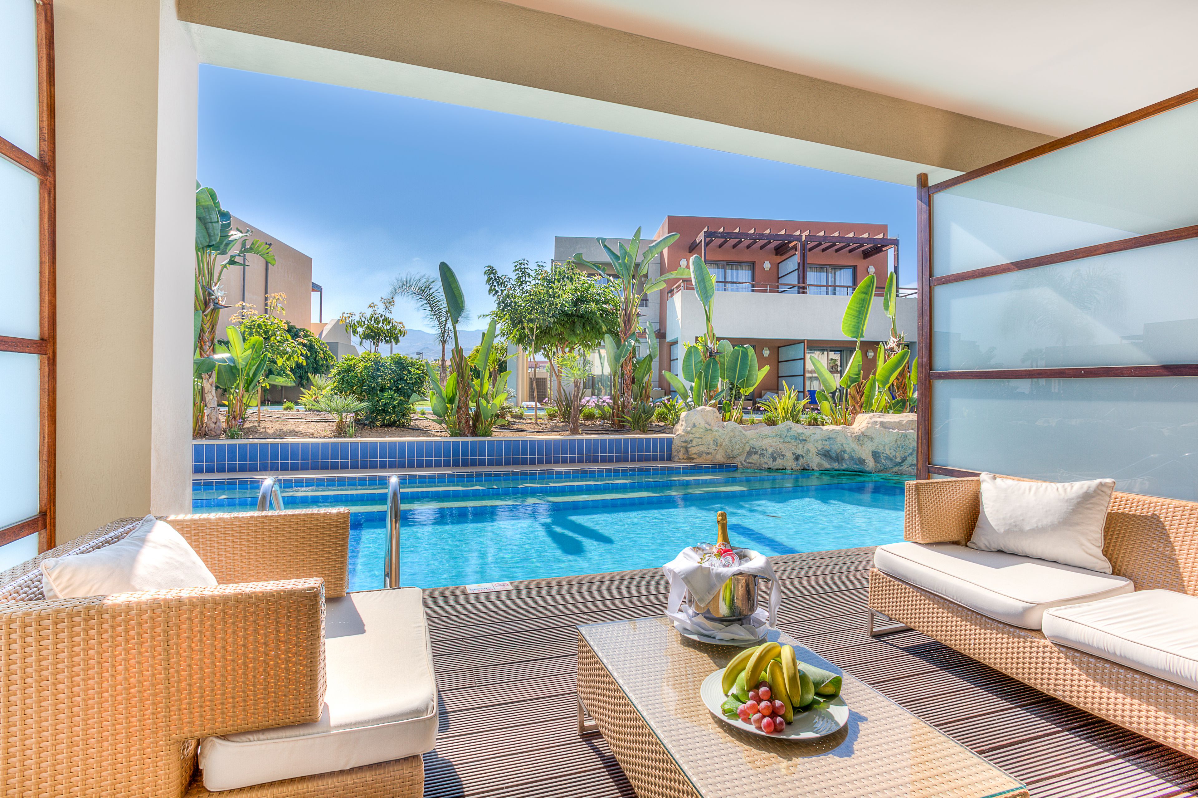 Zona de piscina con mobiliario de mimbre, una botella de champán y una bandeja de fruta.