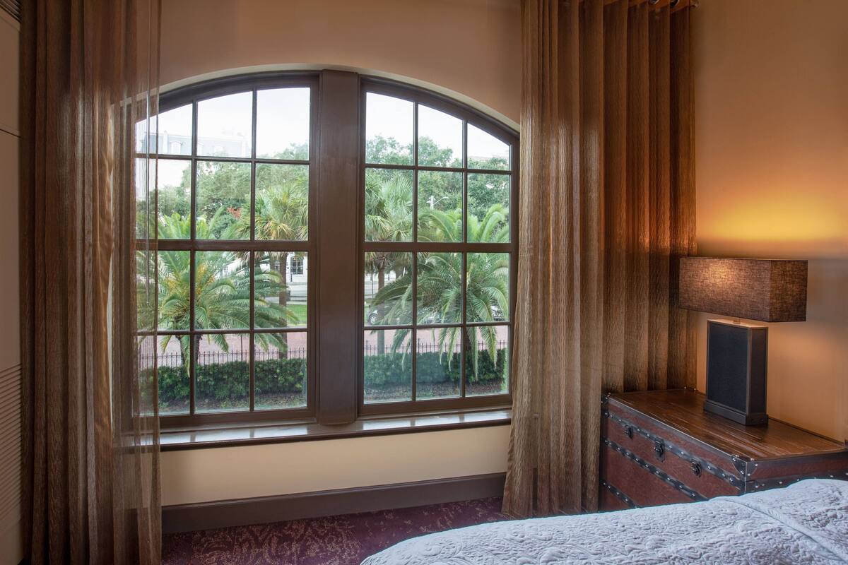 Ein Hotelzimmer mit einem Bogenfenster, einem hölzernen Schminktisch, einer Lampe und einem Bett mit gemustertem Überwurf.