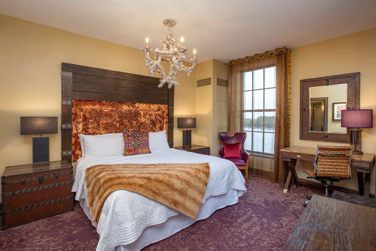 Ein Hotelzimmer mit einem großen Bett, einem hölzernen Kopfteil, einer Chandelüster, einem Schreibtisch mit Stuhl, einem Spiegel und einem fenster mit Vorhängen.