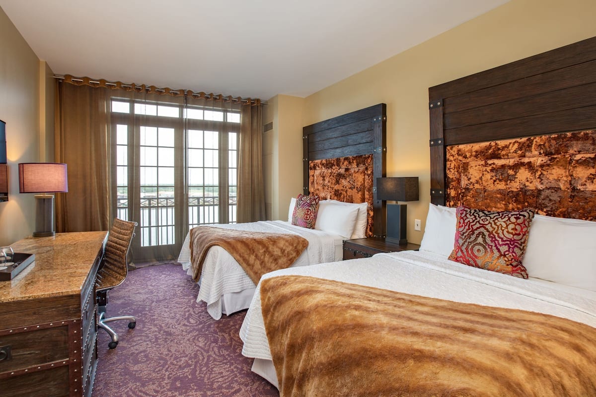 Ein Hotelzimmer mit zwei Betten, einem Schreibtisch und einem Stuhl. Ein großes Fenster mit Vorhängen, und das Zimmer hat ein Holz-Kopfteil.