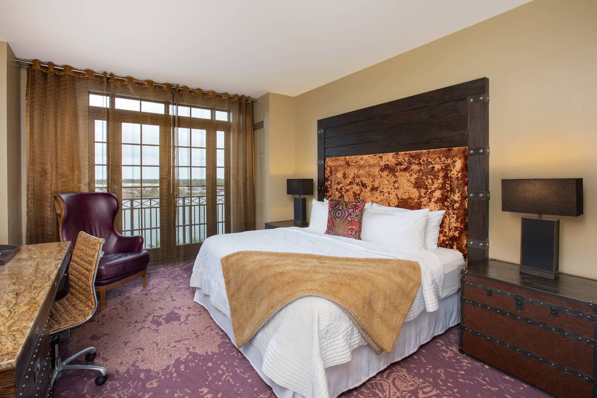 Ein Hotelzimmer mit einem großen Bett, einem Schreibtisch, einem Stuhl, einem Fenster mit Vorhängen und Blick ins Freie.