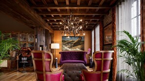 Ein Zimmer mit Holzbalkendecke, einer Kristalllüster und einem violetten Sofa sowie zwei roten Sesseln.