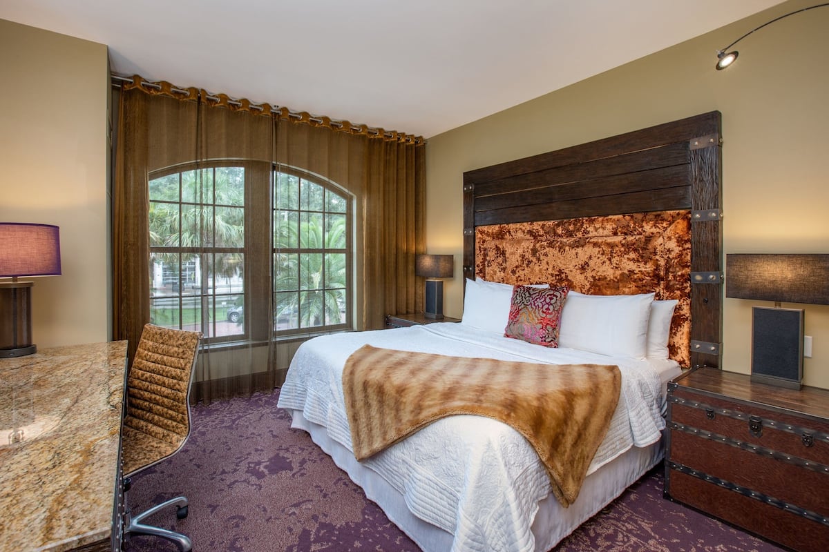 Ein Hotelzimmer mit einem großen Bett, einem Schreibtisch und einem Stuhl. Ein Fenster mit Vorhängen, eine Lampe und ein Koffer auf dem Schminktisch sind zu sehen.