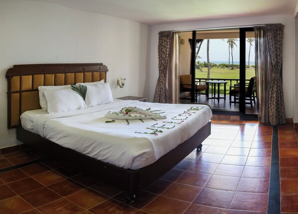 A hotel room with a large bed, a nightstand, a wall-mounted lamp, and a view of the outdoor patio with a table and chairs. - VGP Golden Beach Resort (Chennai)