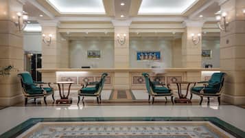A lobby with a reception desk, green upholstered chairs, a patterned carpet, and a tiled floor.