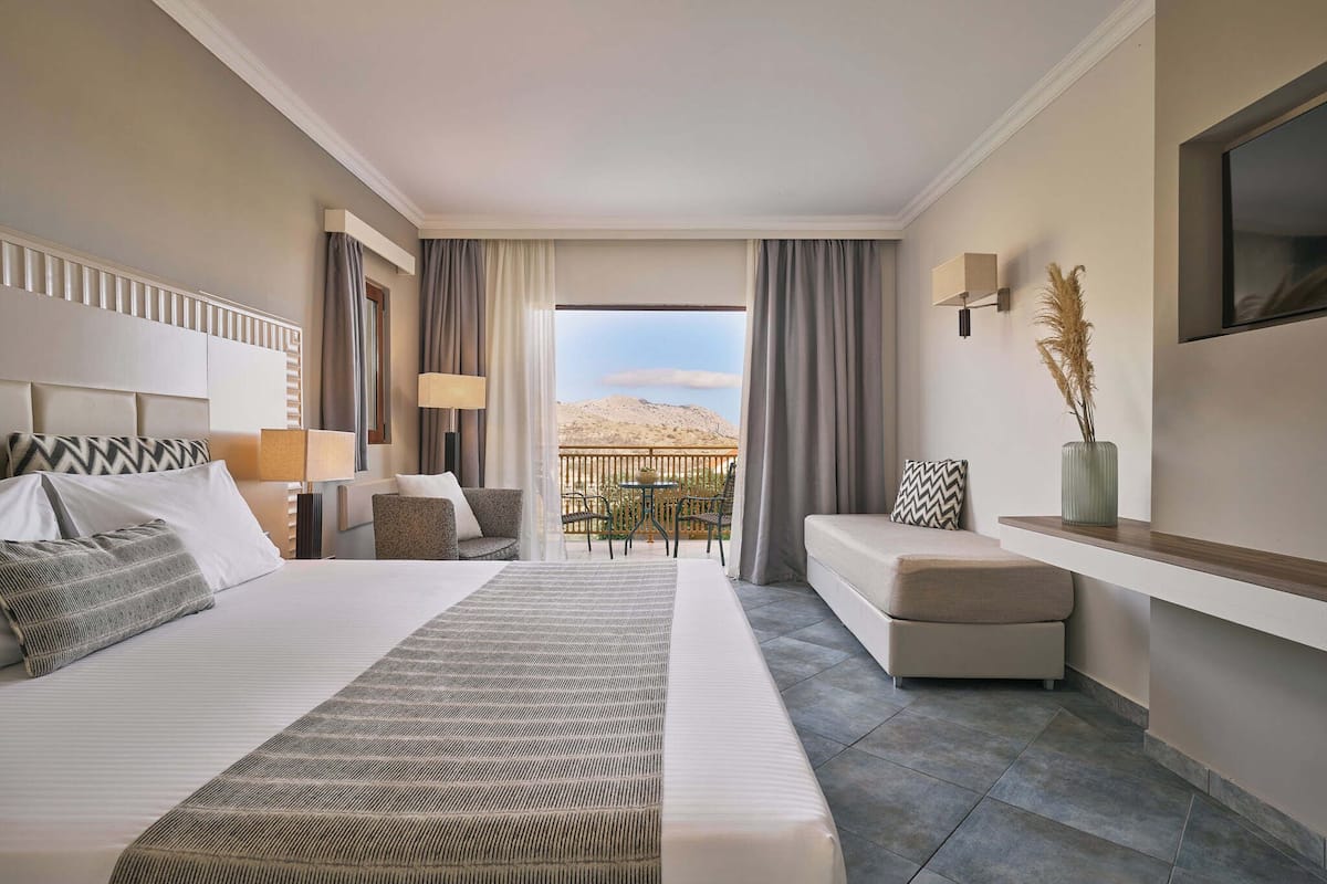 A hotel room with a large bed, a sofa, a small table, and a balcony with a view of the desert.