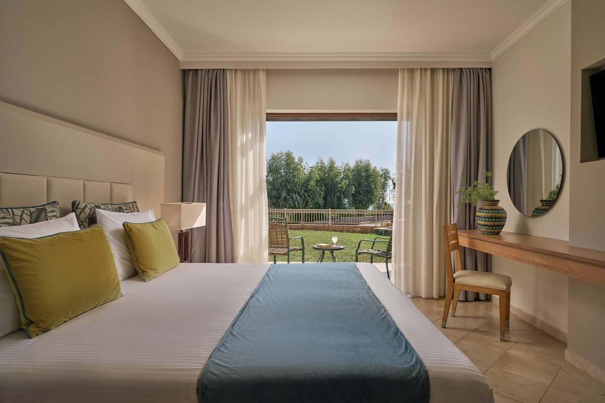 A hotel room with a large bed, a desk, a chair, a small table, and a view of the outdoors.