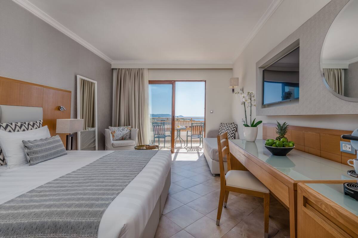 A hotel room with a bed, a desk, a chair, a small table, a bowl of fruit, a potted plant, and a view of the outdoors.