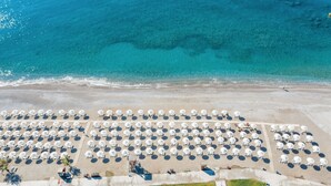 An aerial view of a beach with rows of white umbrellas and beach chairs, a sandy shore, and clear blue water. - Lindos Imperial Resort & Spa (Rhodes)