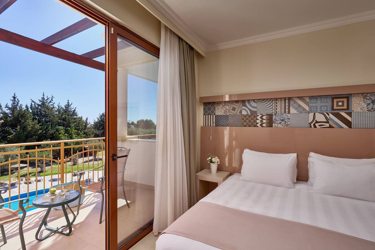A hotel room with a bed, a small table, and a view of a pool and trees.
