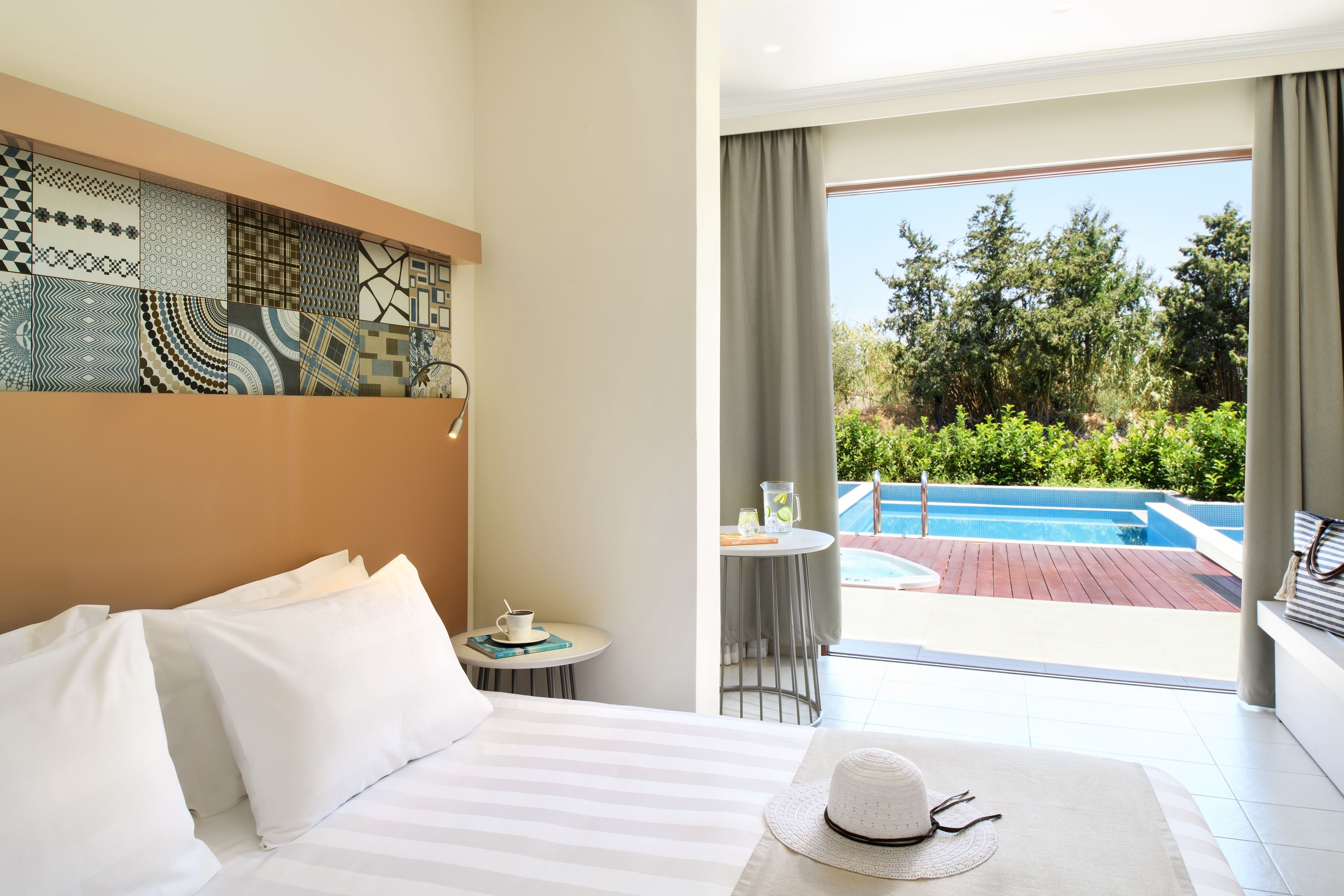 A hotel room with a bed, a small table with a cup and saucer, a hat on the bed, and a view of a swimming pool and greenery outside.