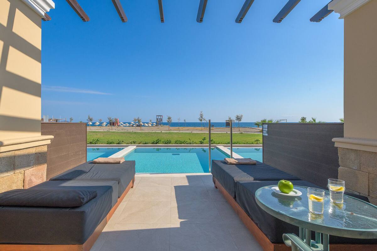 A poolside area with two lounge chairs, a glass table with a green apple and two glasses, and a view of a pool and beach.