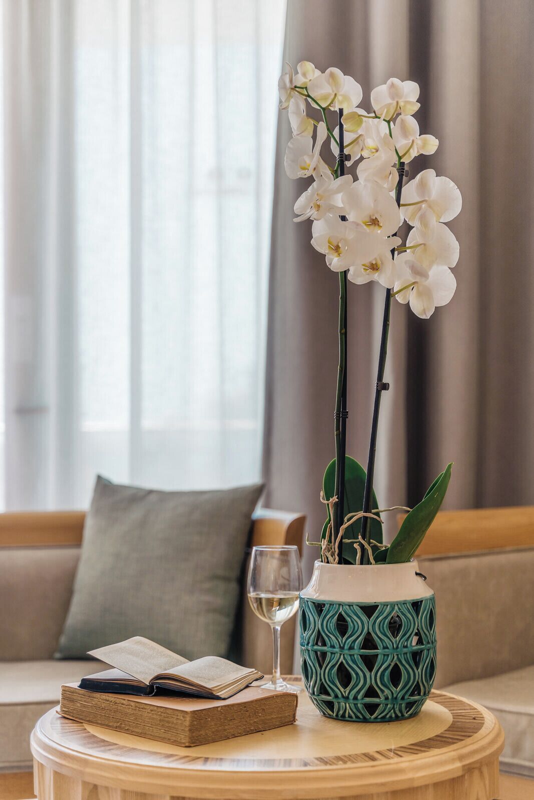 A round wooden table with a vase of white orchids, a book, and a glass of wine.