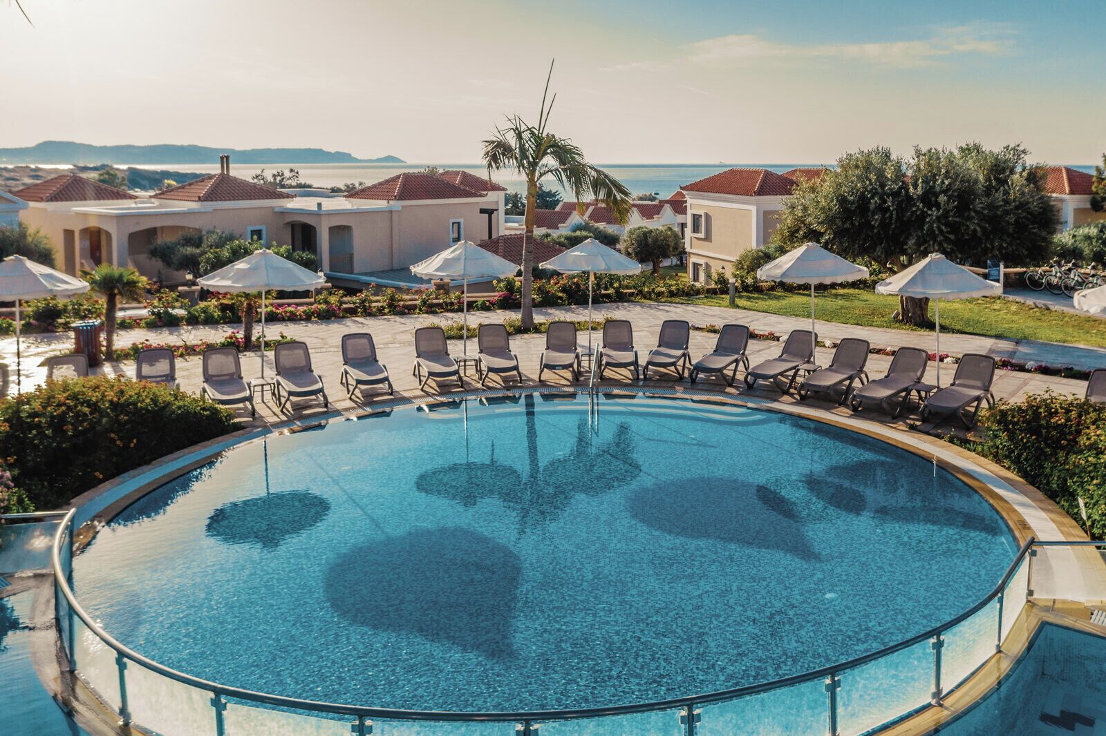 Ein runder Pool mit Liegestühlen und Sonnenschirmen, umgeben von Gebäuden mit roten Ziegeldächern und Blick aufs Meer.