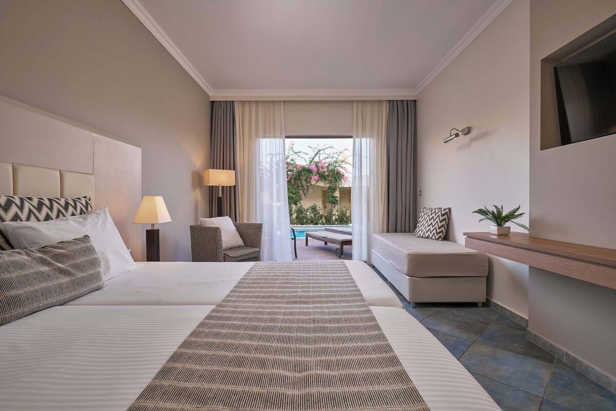 A hotel room with a large bed, a sofa, a small table, and a view of an outdoor pool area.