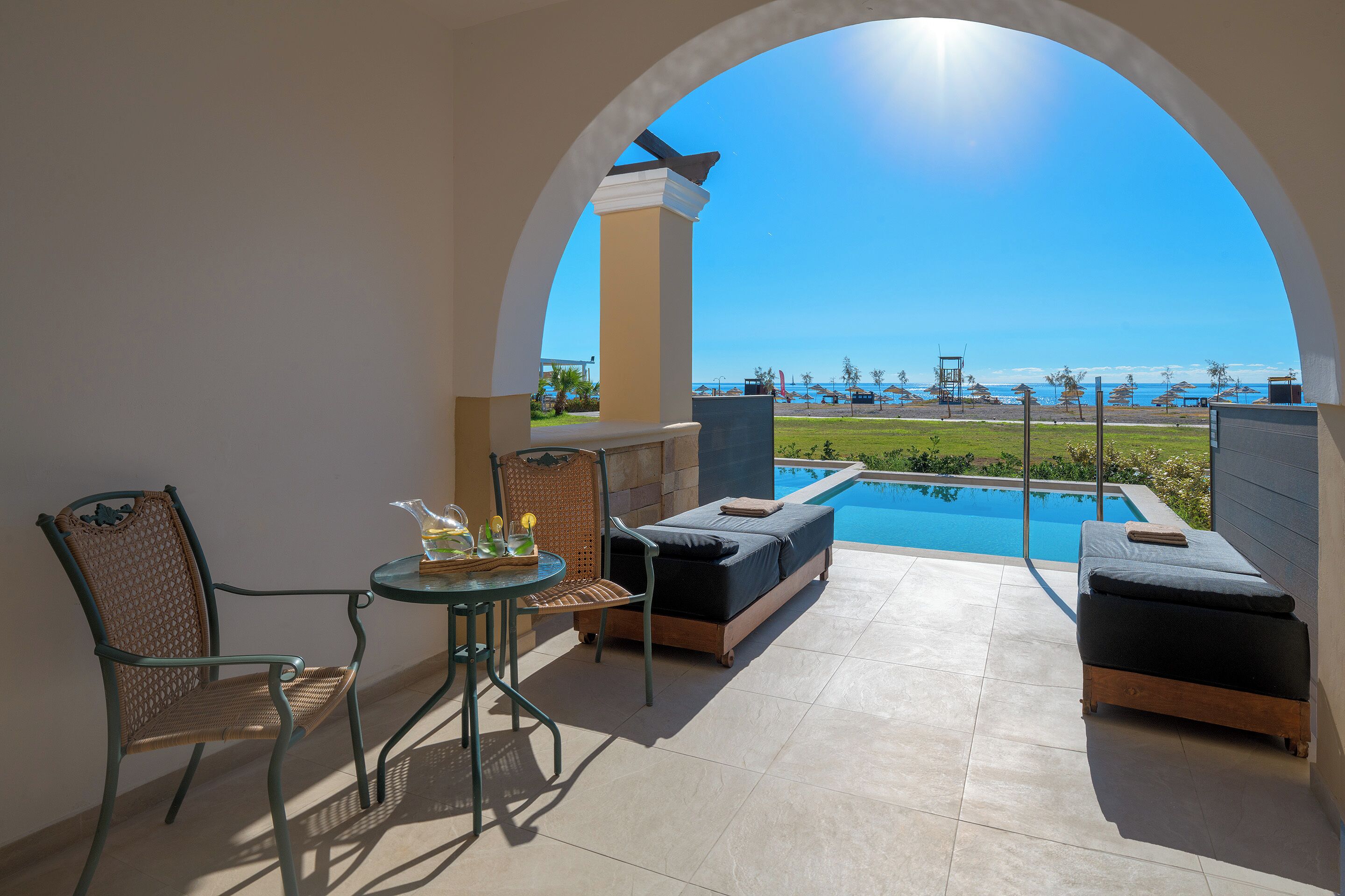 A sunlit patio with a pool, a table with chairs, and a view of the beach.