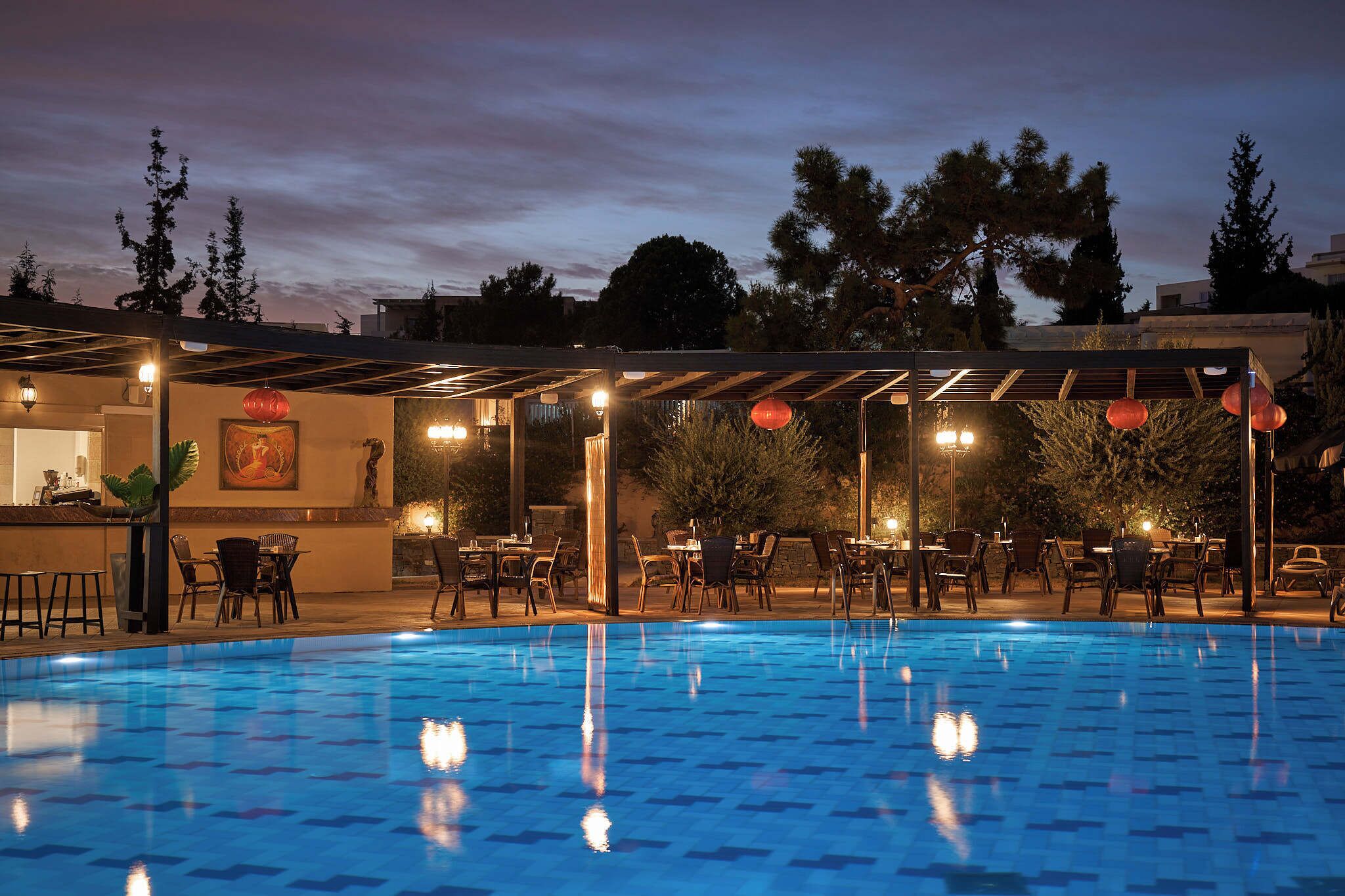 An outdoor swimming pool with a tiled deck, surrounded by tables and chairs, and a covered seating area with string lights.