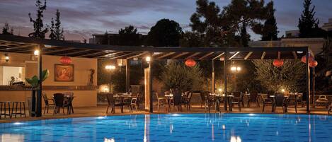 An outdoor swimming pool with a tiled deck, surrounded by tables and chairs, and a covered seating area with string lights.