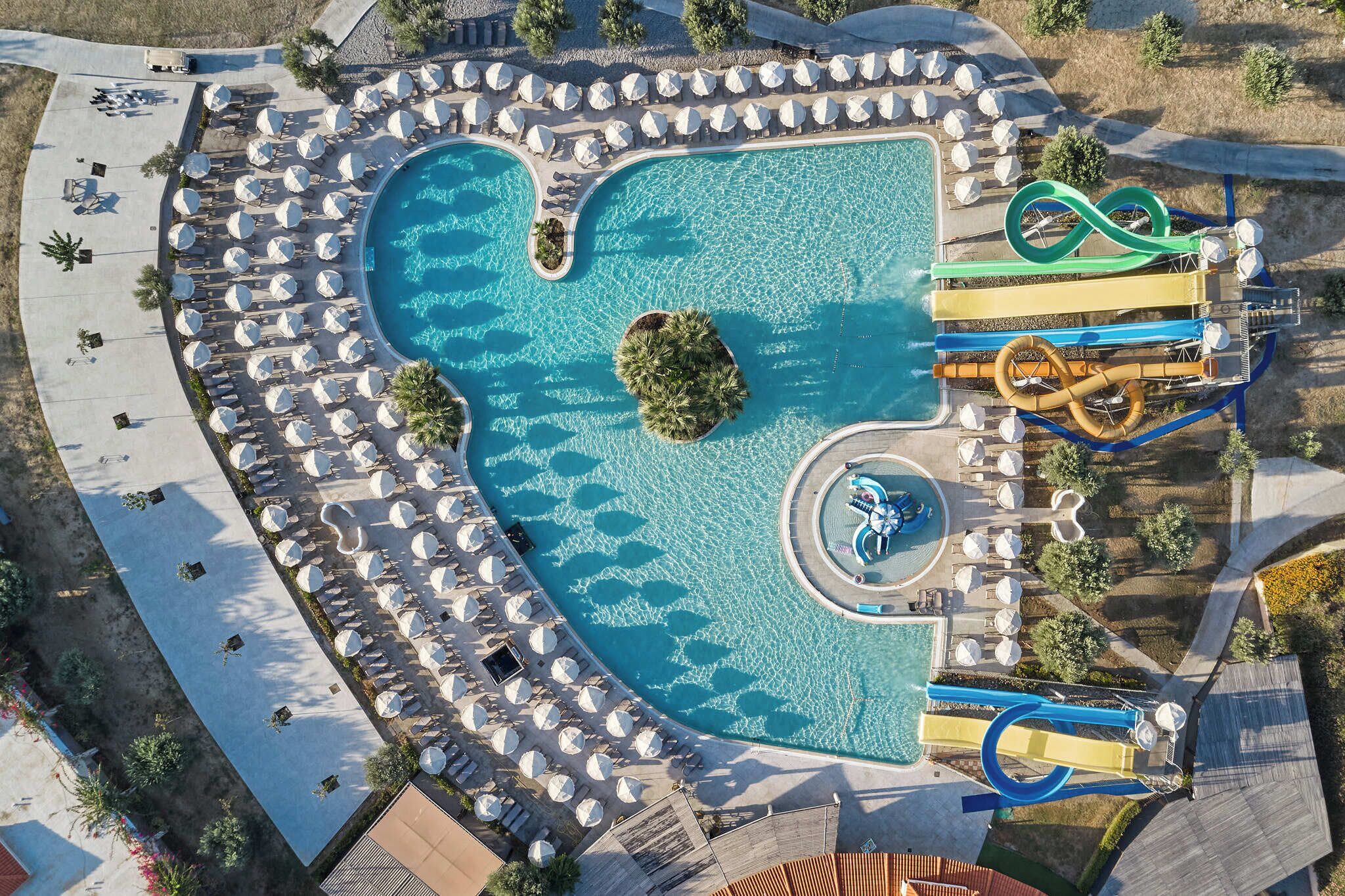 An aerial view of a resort with a large swimming pool, multiple water slides, and sun loungers arranged around the pool area.