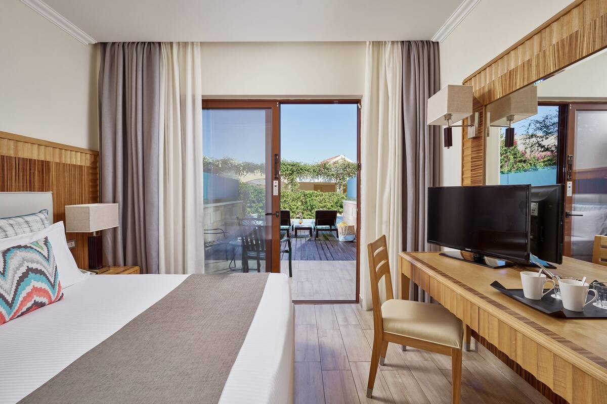 A hotel room with a bed, a desk with a TV, a chair, and a balcony with a table and chairs.