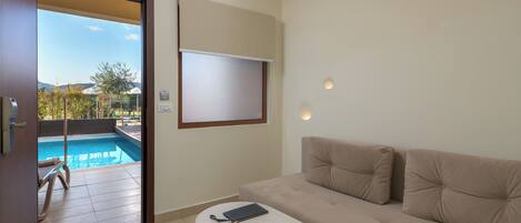 Un salon moderne avec un canapé beige, une petite table ronde sur laquelle se trouvent un livre et des lunettes, et une vue sur une piscine par une porte ouverte.