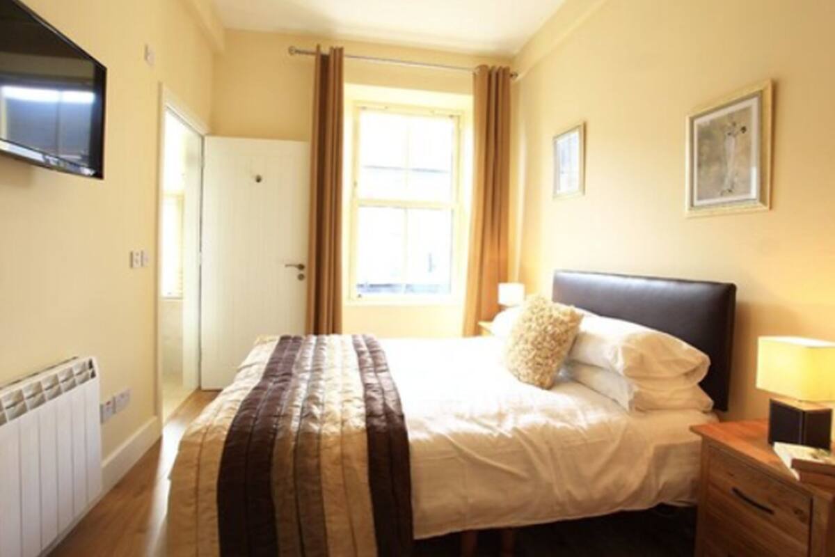 Een slaapkamer met een bed, een nachtkastje, een lamp, een raam met gordijnen, een deur en een aan de muur bevestigde televisie.